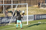 A soccer match scene with two players in focus near the goal. One player, wearing black, is in mid-air reaching to stop or catch the ball, while the other player, wearing teal shorts and black top, is positioned under the ball, looking up at it. The goal net and a fence are visible in the background, with a few spectators standing near the sidelines.