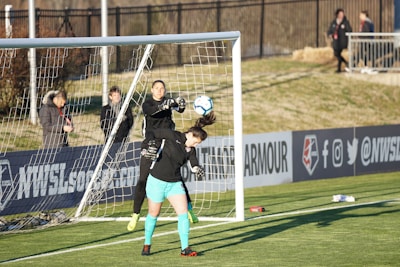 A soccer match scene with two players in focus near the goal. One player, wearing black, is in mid-air reaching to stop or catch the ball, while the other player, wearing teal shorts and black top, is positioned under the ball, looking up at it. The goal net and a fence are visible in the background, with a few spectators standing near the sidelines.