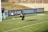 A goalkeeper diving to catch a soccer ball near the goalpost on a grassy field. Another player stands nearby observing the action, with visible advertisements and banners on the fence behind them.