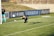 A goalkeeper diving to catch a soccer ball near the goalpost on a grassy field. Another player stands nearby observing the action, with visible advertisements and banners on the fence behind them.