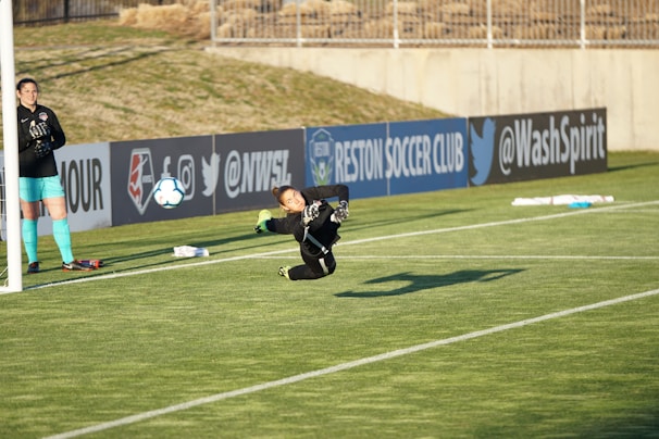 A goalkeeper diving to catch a soccer ball near the goalpost on a grassy field. Another player stands nearby observing the action, with visible advertisements and banners on the fence behind them.
