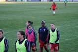 A group of female soccer players wearing training vests and sports attire walking on a green soccer field. Some players are wearing pink vests while others are in neon green. They are participating in a training session, with one player slightly in the background. The field is large and appears well-maintained, bordered in the distance by advertising boards.
