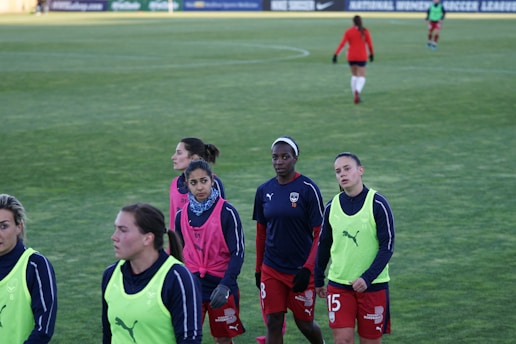 A group of female soccer players wearing training vests and sports attire walking on a green soccer field. Some players are wearing pink vests while others are in neon green. They are participating in a training session, with one player slightly in the background. The field is large and appears well-maintained, bordered in the distance by advertising boards.