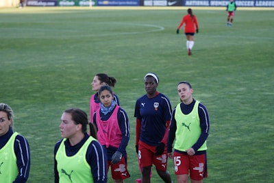 A group of female soccer players wearing training vests and sports attire walking on a green soccer field. Some players are wearing pink vests while others are in neon green. They are participating in a training session, with one player slightly in the background. The field is large and appears well-maintained, bordered in the distance by advertising boards.