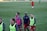 A group of female soccer players wearing training vests and sports attire walking on a green soccer field. Some players are wearing pink vests while others are in neon green. They are participating in a training session, with one player slightly in the background. The field is large and appears well-maintained, bordered in the distance by advertising boards.