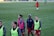 A group of female soccer players wearing training vests and sports attire walking on a green soccer field. Some players are wearing pink vests while others are in neon green. They are participating in a training session, with one player slightly in the background. The field is large and appears well-maintained, bordered in the distance by advertising boards.