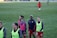 A group of female soccer players wearing training vests and sports attire walking on a green soccer field. Some players are wearing pink vests while others are in neon green. They are participating in a training session, with one player slightly in the background. The field is large and appears well-maintained, bordered in the distance by advertising boards.