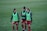 Three soccer players are walking on a grass field, wearing red shorts, blue long-sleeve shirts, and green training bibs with a Puma logo. They appear to be in discussion or conversation, showing a relaxed and friendly demeanor.