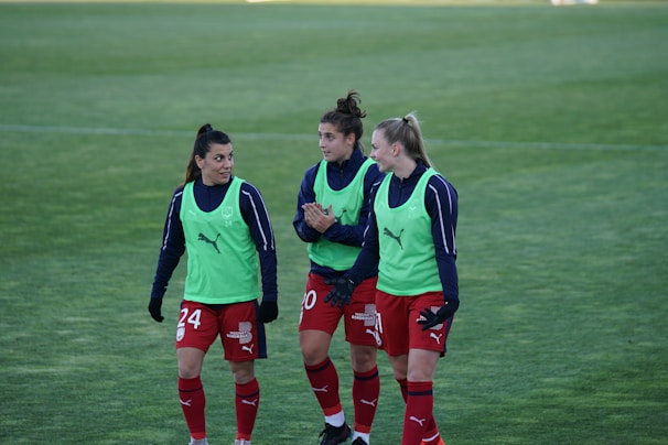 Three soccer players are walking on a grass field, wearing red shorts, blue long-sleeve shirts, and green training bibs with a Puma logo. They appear to be in discussion or conversation, showing a relaxed and friendly demeanor.
