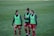 Three soccer players are walking on a grass field, wearing red shorts, blue long-sleeve shirts, and green training bibs with a Puma logo. They appear to be in discussion or conversation, showing a relaxed and friendly demeanor.