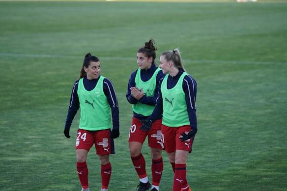 Three soccer players are walking on a grass field, wearing red shorts, blue long-sleeve shirts, and green training bibs with a Puma logo. They appear to be in discussion or conversation, showing a relaxed and friendly demeanor.