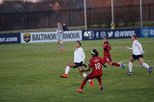 A women's soccer match is taking place on a green field. Several players in red and white uniforms are focused on a soccer ball in mid-air. The players show various athletic postures, indicating intense action and engagement. In the background, there are advertising boards and a fence separating the field from the trees beyond.