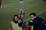 A group of individuals on a sports field are wearing green vests over red attire. One person is holding a tablet and seems to be discussing or showing something to the others. The atmosphere suggests a sports event, likely soccer, indicated by their clothing and the setting. The background is a grassy field.
