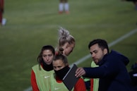 A group of individuals on a sports field are wearing green vests over red attire. One person is holding a tablet and seems to be discussing or showing something to the others. The atmosphere suggests a sports event, likely soccer, indicated by their clothing and the setting. The background is a grassy field.