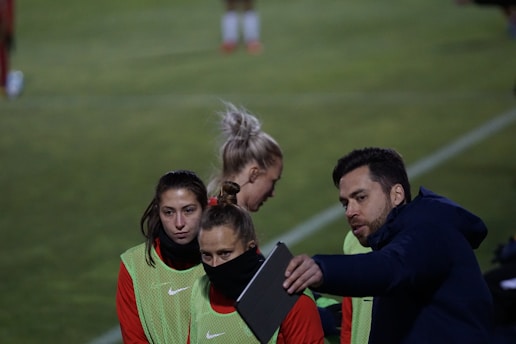 A group of individuals on a sports field are wearing green vests over red attire. One person is holding a tablet and seems to be discussing or showing something to the others. The atmosphere suggests a sports event, likely soccer, indicated by their clothing and the setting. The background is a grassy field.