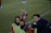 A group of individuals on a sports field are wearing green vests over red attire. One person is holding a tablet and seems to be discussing or showing something to the others. The atmosphere suggests a sports event, likely soccer, indicated by their clothing and the setting. The background is a grassy field.