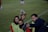 A group of individuals on a sports field are wearing green vests over red attire. One person is holding a tablet and seems to be discussing or showing something to the others. The atmosphere suggests a sports event, likely soccer, indicated by their clothing and the setting. The background is a grassy field.