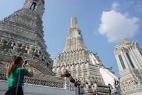 An intricately detailed temple with ornate carvings and patterns on its towers, highlighting its architectural beauty under a clear blue sky. A person is capturing the scene with a camera, indicating awe and admiration for the structure.