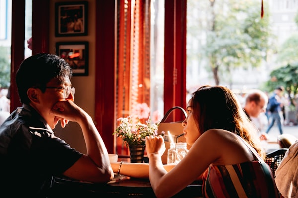 Couple on a date sitting at a table with flowers, drinks in front of a window with people passing outside.