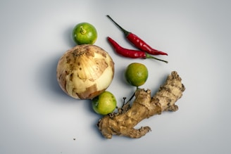 A collection of fresh ingredients including a large yellow-brown onion, a piece of ginger root, two red chili peppers, and three green limes. They are arranged on a smooth, light grey surface.