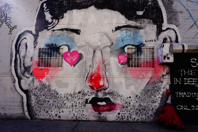 A street art mural painted on a brick wall depicting a stylized face with exaggerated features. The face has large, expressive eyes with heart shapes incorporated into the design. The nose is prominently painted in a bright red color, and a vibrant pink is used for the cheeks. The entire mural is marked with dark outlines, and there are two small barred windows positioned as if they are the character's eyes.
