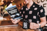 A person is reading a book titled 'Expert Secrets' in a warm, cozy setting. They are holding a black mug with '80/20' printed on it. A closed laptop rests on the wooden table next to them. The person is wearing a patterned shirt and a white watch. Soft orange and yellow lights create a relaxed ambiance.