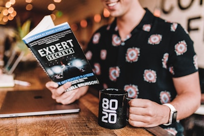 A person is reading a book titled 'Expert Secrets' in a warm, cozy setting. They are holding a black mug with '80/20' printed on it. A closed laptop rests on the wooden table next to them. The person is wearing a patterned shirt and a white watch. Soft orange and yellow lights create a relaxed ambiance.