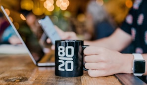 Entrepreneur holding a 'Boss That Brew' mug while working on a laptop in a sunlit modern workspace with leather and chalkboard decor