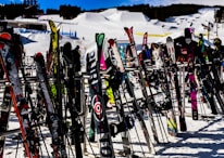 Numerous skis and snowboards are standing upright on racks, set against a snowy backdrop of a ski resort with slopes and structures in the distance.