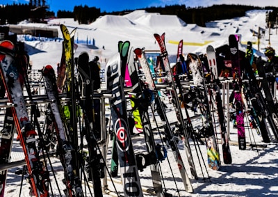 Numerous skis and snowboards are standing upright on racks, set against a snowy backdrop of a ski resort with slopes and structures in the distance.
