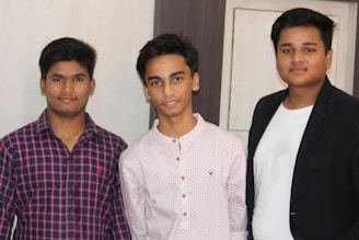 Three young men standing side by side indoors. They are dressed in casual attire, with one wearing a checkered shirt, another in a patterned shirt, and the third in a white shirt with a dark blazer. The background is neutral, featuring a wall with a decorative panel.