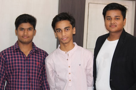 Three young men standing side by side indoors. They are dressed in casual attire, with one wearing a checkered shirt, another in a patterned shirt, and the third in a white shirt with a dark blazer. The background is neutral, featuring a wall with a decorative panel.