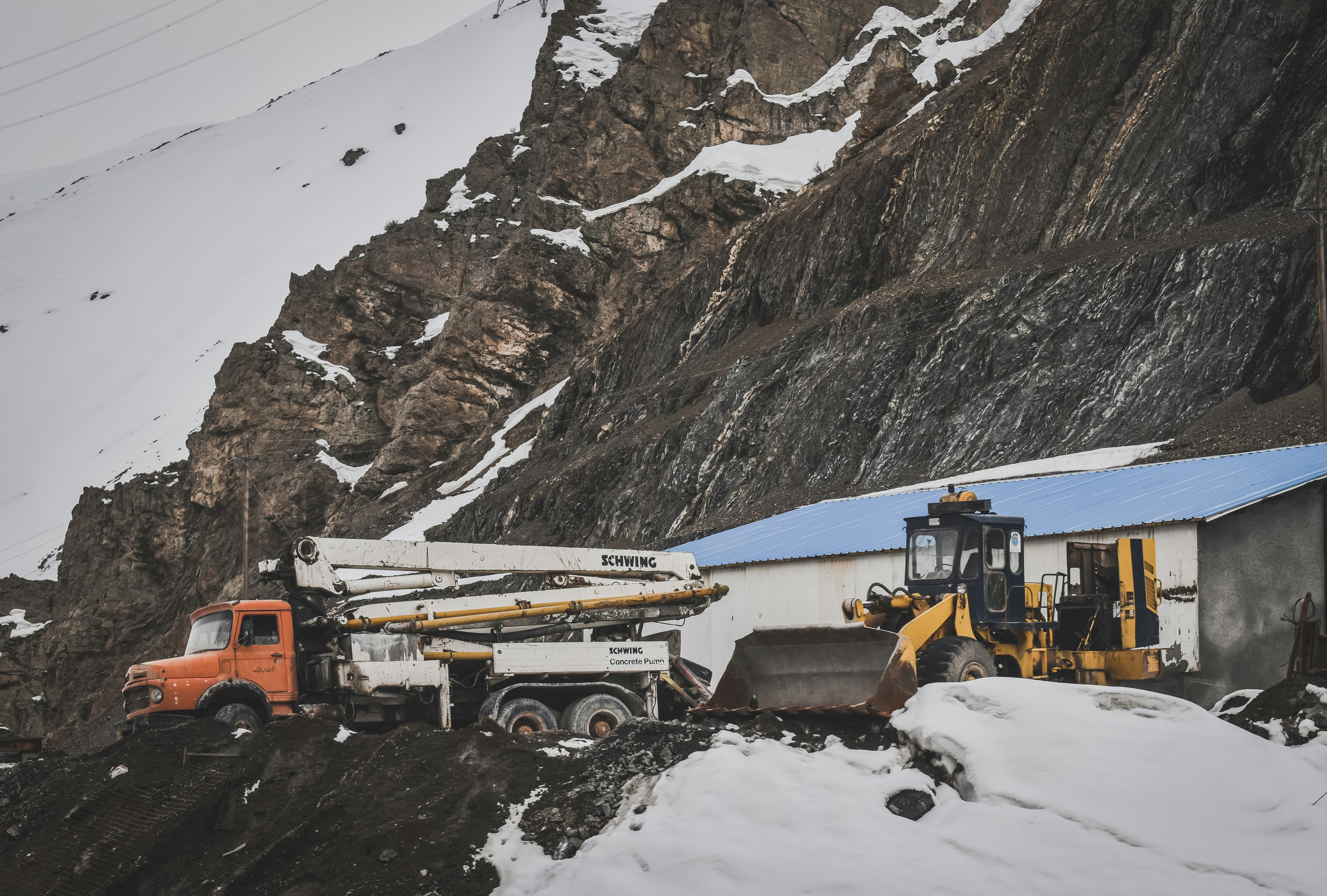Snow-covered remote worksite with an orange truck and a yellow excavator beside a blue-roofed building on a rocky slope.