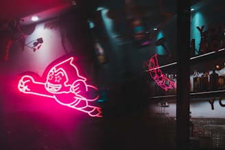 A vibrant pink neon light in the shape of a cartoon character with starry eyes and a smiling expression, positioned as if flying forward. The background is a dimly lit bar area with bottles and glassware, providing a contrast between the colorful neon and the darker surroundings. Reflections of the neon light are visible on a glass surface.