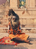 An elderly man with long, tied-up hair and a large beard sits cross-legged on a colorful mat. He is adorned with layers of bead necklaces and wears an orange garment. His hands rest on his lap, holding a small stick. The background features ancient stone steps and a carved wall with an ornate sculpture.