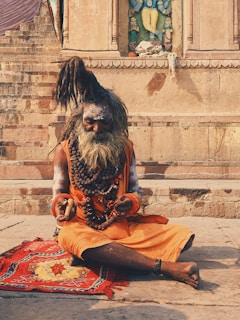 An elderly man with long, tied-up hair and a large beard sits cross-legged on a colorful mat. He is adorned with layers of bead necklaces and wears an orange garment. His hands rest on his lap, holding a small stick. The background features ancient stone steps and a carved wall with an ornate sculpture.