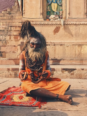 An elderly man with long, tied-up hair and a large beard sits cross-legged on a colorful mat. He is adorned with layers of bead necklaces and wears an orange garment. His hands rest on his lap, holding a small stick. The background features ancient stone steps and a carved wall with an ornate sculpture.