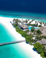 A tropical island resort featuring white sandy beaches and turquoise waters. Palm trees line the shore, and a wooden pier extends into the ocean. Thatched-roof bungalows are nestled among lush greenery, with a clear blue swimming pool visible near the center of the image.