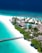 A tropical island resort featuring white sandy beaches and turquoise waters. Palm trees line the shore, and a wooden pier extends into the ocean. Thatched-roof bungalows are nestled among lush greenery, with a clear blue swimming pool visible near the center of the image.