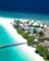 A tropical island resort featuring white sandy beaches and turquoise waters. Palm trees line the shore, and a wooden pier extends into the ocean. Thatched-roof bungalows are nestled among lush greenery, with a clear blue swimming pool visible near the center of the image.