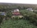 An aerial view of a lush, green landscape featuring multiple houses surrounded by dense vegetation and plantations. The central focus is a large house with a distinctive red roof, surrounded by banana plants and various trees. Other houses are scattered throughout the area, immersed in the verdant environment.