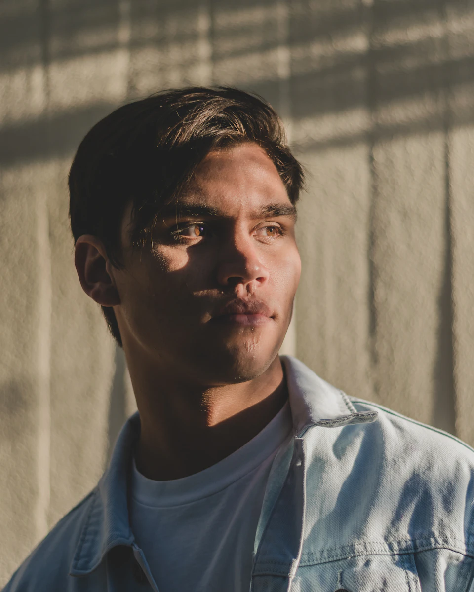 The image shows a young man with short dark hair wearing a white t-shirt and denim jacket, standing against a light beige wall. The man's gaze is directed off to the side, suggesting he may be lost in thought or observing something outside of the frame.