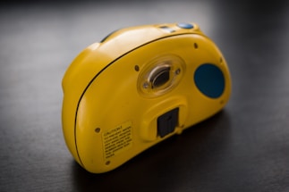 A yellow plastic device resembling a camera is placed on a black surface. It has a lens area positioned centrally and a contrasting blue circular design on one side. The device has a label with cautionary text on the back.