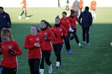 A group of people are engaged in a soccer practice session on a grassy field. They are wearing matching red jerseys and black pants, some with white socks and gloves. The participants seem focused and are jogging or walking, possibly warming up.