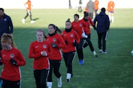 A group of people are engaged in a soccer practice session on a grassy field. They are wearing matching red jerseys and black pants, some with white socks and gloves. The participants seem focused and are jogging or walking, possibly warming up.