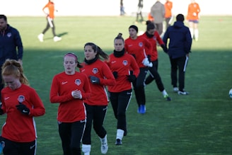 A group of people are engaged in a soccer practice session on a grassy field. They are wearing matching red jerseys and black pants, some with white socks and gloves. The participants seem focused and are jogging or walking, possibly warming up.