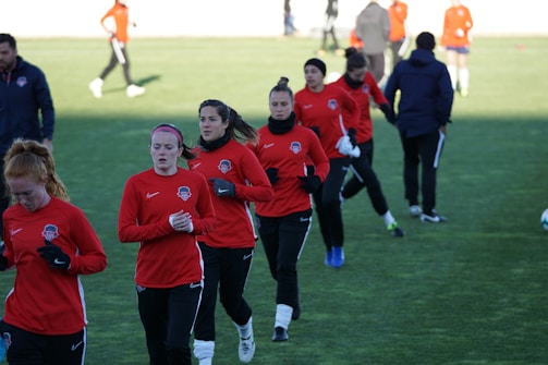 A group of people are engaged in a soccer practice session on a grassy field. They are wearing matching red jerseys and black pants, some with white socks and gloves. The participants seem focused and are jogging or walking, possibly warming up.