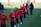 A group of people are engaged in a soccer practice session on a grassy field. They are wearing matching red jerseys and black pants, some with white socks and gloves. The participants seem focused and are jogging or walking, possibly warming up.