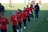 A group of people are engaged in a soccer practice session on a grassy field. They are wearing matching red jerseys and black pants, some with white socks and gloves. The participants seem focused and are jogging or walking, possibly warming up.