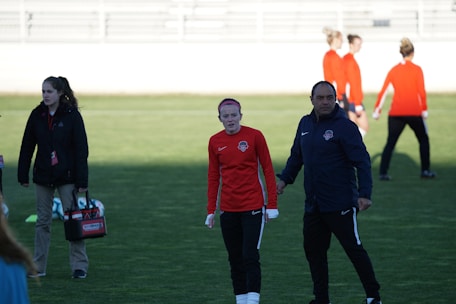 A group of people are gathered on a sports field. Some individuals are dressed in athletic gear, including a woman in a red sports jersey and a man beside her in a blue jacket, appearing to be in conversation. Additional people in similar athletic clothing are visible in the background.